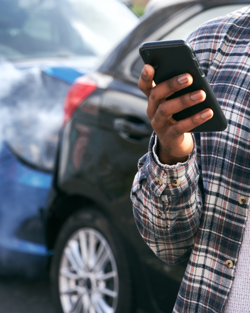 Persona marcando un teléfono celular frente a un choque de vehículos